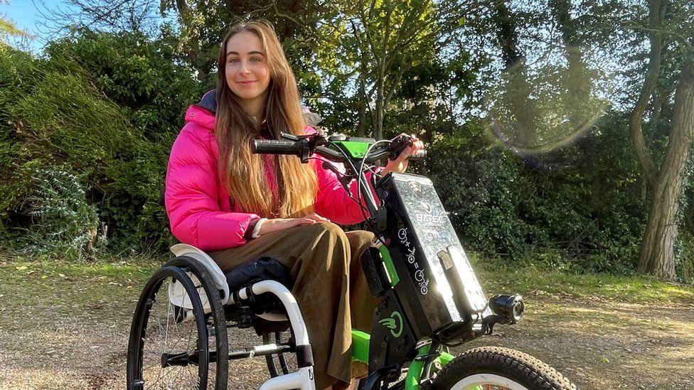 Bethany Handley sitting in her wheelchair in a park. She is a young female wearing a bright pink coat.
